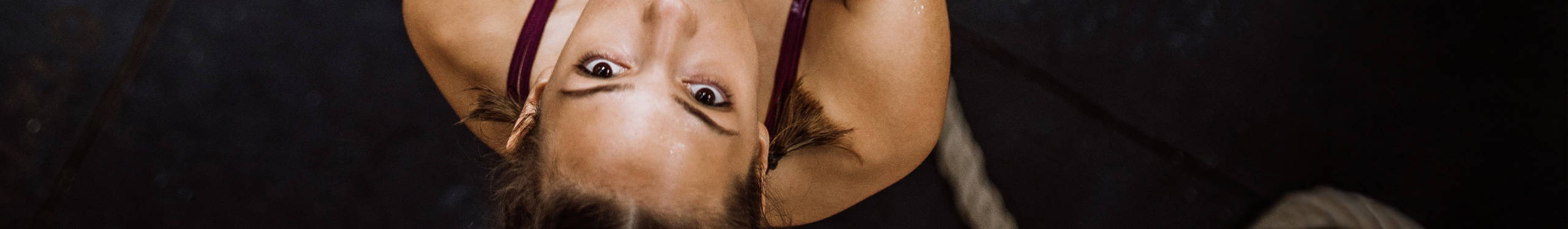 Eine Person hängt kopfüber an einem Seil, das in einem dunklen Raum befestigt ist. Sie trägt ein ärmelloses Oberteil. Die Szene vermittelt ein Gefühl von Konzentration und Balance.