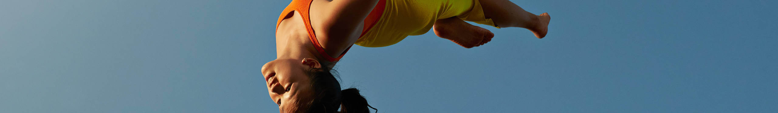 Eine Person in einem gelben und orangefarbenen Sportoutfit führt einen Salto in der Luft aus, vor einem klaren blauen Himmel. Die Szene vermittelt Dynamik und Bewegungsfreiheit.