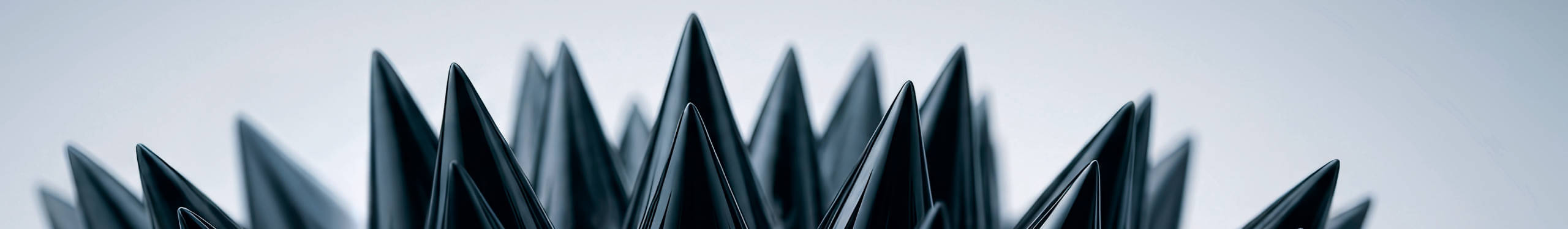 Eine Nahaufnahme von spitzen, schwarzen Strukturen, die an eine Reihe von Kegeln oder Stacheln erinnern. Die Formen sind symmetrisch angeordnet und heben sich vor einem unscharfen, hellen Hintergrund ab. Die Anordnung und das Material der Strukturen erzeugen einen futuristischen und abstrakten Eindruck.